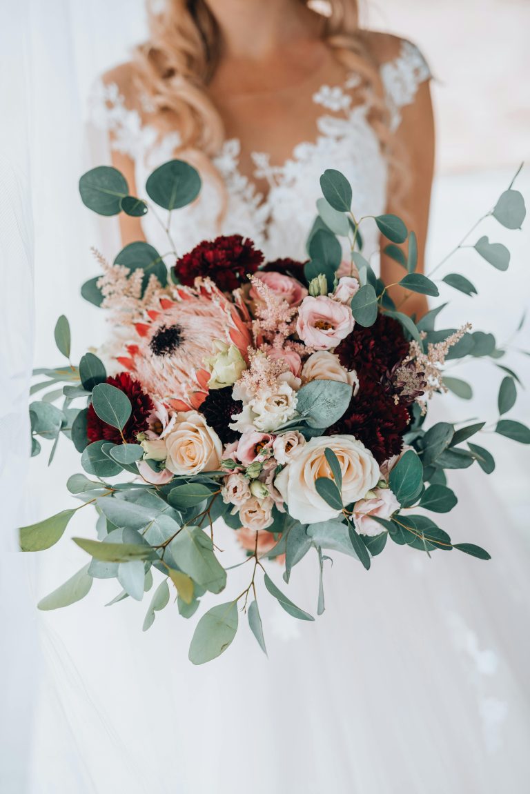 Bride in white lace gown showcasing a beautiful bouquet with pink and red flowers.